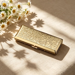 May include: A rectangular gold-coloured metal case featuring an intricate embossed scrollwork pattern across the top surface. The case has a hinged closure mechanism along the side and a smooth, polished finish that reflects light across its surface.