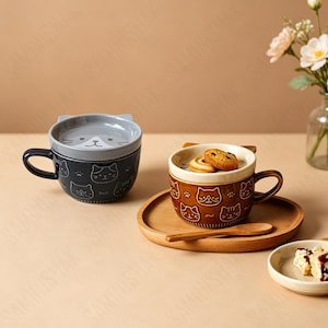 以下が含まれることがあります： 猫の顔の模様と猫耳型の蓋が付いた、ダークグレーとブラウンのセラミック製マグカップ2点。ブラウンのマグカップは木製のトレイに置かれ、木製のスプーンとクッキーが入っています。グレーのマグカップは蓋が閉まった状態です。