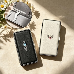 May include: Two rectangular metal cases with textured covers, one black and one white. The black case features a blue eye charm, while the white case has a pink heart with wings. An inset shows the interior with a metal clip engraved with the name William.