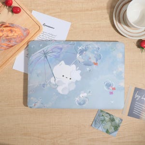 May include: A light blue laptop case featuring a whimsical illustration of a white bear holding an umbrella, floating in a dreamy sky with bubbles. The case has a matte finish and is surrounded by other objects on a wooden surface.