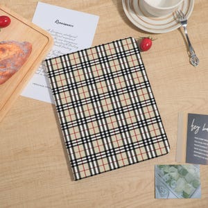 May include: A beige and plaid patterned notebook or tablet cover. The cover features a repeating pattern of black, white, and red lines over a beige background. The notebook is on a wooden surface with other objects.
