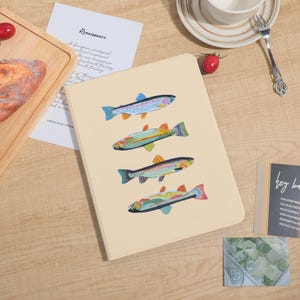 Può includere: Un quaderno beige con illustrazioni di pesci colorati. I pesci sono raffigurati in blu, verde, arancione e giallo. Il quaderno è aperto e i pesci sono disposti verticalmente.