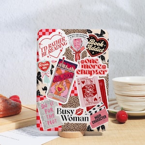 May include: A decorative book cover featuring various stickers with book-related themes. Stickers include phrases like "I'd Rather Be Reading," "Book Addict," and "One More Chapter." The design incorporates a leopard print background and a red and pink color scheme.