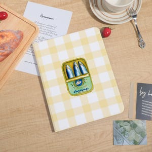 May include: A notebook with a yellow and white gingham pattern. The cover features a sardine can illustration with three fish. The notebook is on a wooden surface with other items.