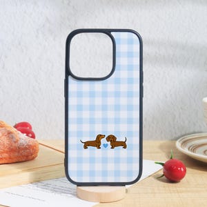 May include: A phone case with a light blue and white gingham check pattern. The design features two brown dachshunds facing each other with a small light blue heart positioned between them. The case has a black border and a camera cutout.