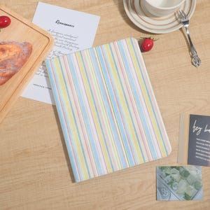 Op de afbeelding: Een gesloten notitieboek met een pastel gestreept patroon in geel, blauw en roze. Het notitieboek ligt op een houten oppervlak met een stuk brood, een tomaat en een kopje koffie.