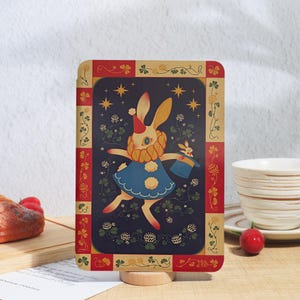 May include: A rectangular card featuring a cream-colored rabbit dressed as a clown in a blue dress with a red hat and yellow ruff. The rabbit holds a blue top hat with a smaller rabbit inside. The background is dark blue with stars and clover plants.