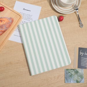 Może przedstawiać: Notatnik z miętowo-zieloną i białą okładką w paski jest centralnym punktem tego zdjęcia. Notatnik leży na drewnianej powierzchni, z kawałkiem chleba, pomidorem oraz filiżanką i spodkiem w pobliżu. Widoczna jest również karta ze słowem "Renaissance".