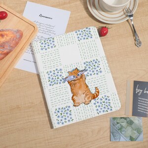 Puede incluir: Un cuaderno blanco con una ilustración de un gato sosteniendo dos peces. El cuaderno tiene un patrón floral con flores azules y guiones verdes. La palabra "Renaissance" está en un papel.
