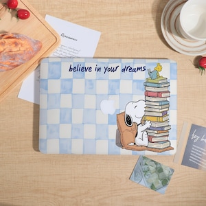May include: A laptop skin featuring a light blue and white checkered pattern with the text "believe in your dreams" and an illustration of Snoopy sitting on a brown cushion next to a tall stack of colorful books with Woodstock on top.