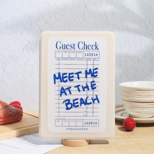 May include: A cream-colored guest check holder with a blue and white design. The text "MEET ME AT THE BEACH" is written in blue. The holder is on a wooden stand, with a cup and cherry in the background.