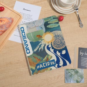 May include: A notebook with a colorful ocean-themed design. The cover features a goldfish in a bowl, a sun, mountains, and the words "OCEANO" and "PACIFIC". The background is a mix of blue, green, and white.