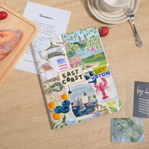 May include: A notebook with a colorful collage design featuring a lighthouse, American flag, lobster, and the words "East Coast" and "Boston". The notebook is open on a wooden table with other items.