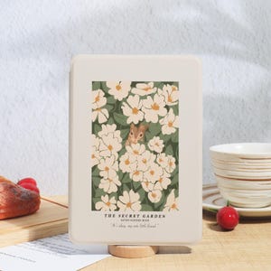 May include: A rectangular, off-white box featuring a floral illustration with a squirrel peeking through white flowers. The text "THE SECRET GARDEN" and "It's okay, my cute little friend." are printed below the image. The box is on a wooden stand.