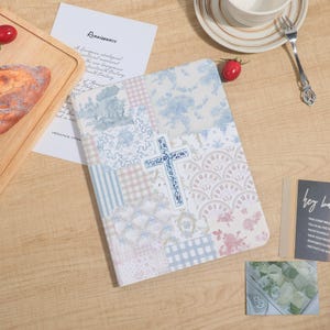 Peut inclure: Un journal décoratif avec un motif patchwork composé de motifs floraux, vichy et toile de Jouy aux couleurs pastel. Une croix bleue est centrée sur la couverture. Le mot "Renaissance" est imprimé sur un papier blanc.