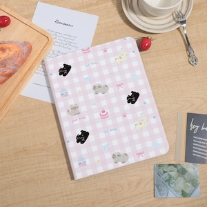 Könnte beinhalten: Ein rosa-weiß kariertes Notizbuch mit Katzenillustrationen, Schleifen und dem Text "Cat Baby" und "pink Daily". Das Notizbuch liegt offen auf einer Holzoberfläche mit anderen Gegenständen.