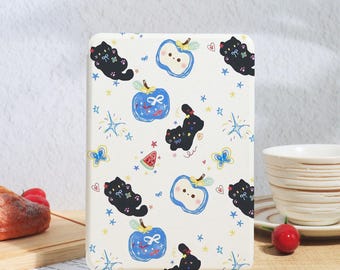 Pode incluir: Uma capa de tablet creme com um padrão repetido de gatos pretos, maçãs azuis, borboletas, estrelas e fatias de melancia. A capa é exibida em um suporte de madeira, com uma xícara e uma cereja no fundo.
