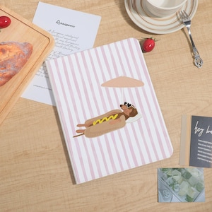 May include: A notebook with a pink and white striped cover featuring an illustration of a dachshund relaxing on a hot dog, wearing sunglasses, and under a parasol. The notebook is on a wooden surface.