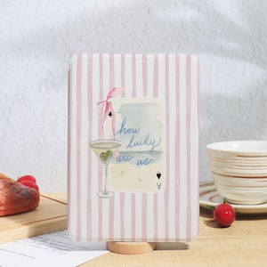 May include: A light pink striped case featuring a watercolour illustration of a martini glass, a pink bow, and the text "how lucky are we." The design includes a playing card and a beach scene.