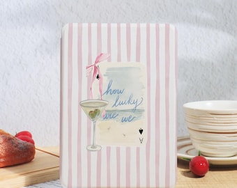 May include: A light pink striped case with a watercolor illustration of a martini glass, a pink bow, and the text "how lucky are we." The design includes a playing card and a beach scene.