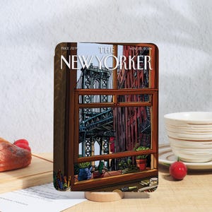 Può includere: Un blocco di legno con un'illustrazione della copertina della rivista New Yorker raffigurante una scena cittadina con un ponte ed edifici. La copertina include il testo "THE NEW YORKER" e la data "NOV 25, 2021".