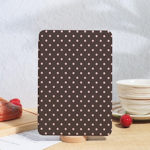 May include: A rectangular brown object featuring a pattern of pink polka dots. The object is presented on a wooden stand. The background includes a white teacup, a plate, and a red cherry.