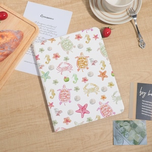 May include: A white notebook with a watercolor pattern of sea turtles, starfish, crabs, seahorses, and flowers. The notebook is open, and a paper with the word "Renaissance" is visible.
