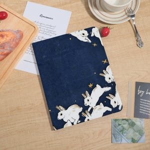 Può includere: Un quaderno blu navy con un motivo di conigli bianchi e stelle dorate. Il quaderno è aperto su un tavolo di legno con pane, un pomodoro e una tazza di caffè.