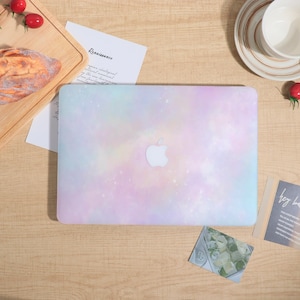 Op de afbeelding: Een pastelkleurige laptophoes met een galaxy-achtig ontwerp. De hoes heeft een zachte mix van roze, blauw en geel, met een wit Apple-logo in het midden. Op een houten tafel staan ook een kopje, een schotel en kaarten.