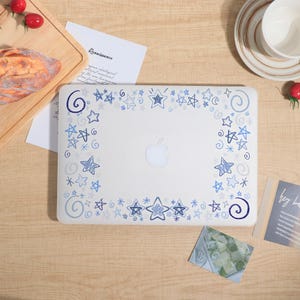 May include: A white laptop with a blue star and swirl design around the edges. The Apple logo is visible in the center. The laptop is on a wooden surface with other items, including a cutting board and a cup.