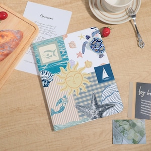 Può includere: Un taccuino rettangolare con una copertina in stile collage che presenta illustrazioni di una tartaruga marina, un sole, una stella marina, una sirena, conchiglie e una barca a vela. Il design incorpora vari motivi, tra cui righe e quadretti.