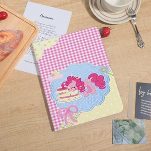 May include: A rectangular notebook featuring a pink and white gingham pattern with a central blue scalloped frame. Inside the frame, a pink pony character is depicted eating a layered cake. The notebook has yellow corner accents with stars.