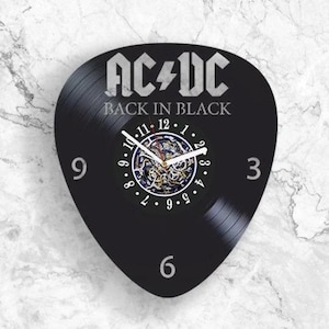 Op de afbeelding: Een wandklok in de vorm van een plectrum, gemaakt van een zwarte vinylplaat. De wijzerplaat toont het AC/DC-logo en de tekst Back in Black in het wit. De klok heeft witte wijzers, de cijfers 3, 6, 9 en 12, en een zichtbaar tandwielmechanisme.