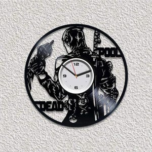 Puede incluir: Un reloj de pared circular hecho de un disco de vinilo negro con una silueta recortada de un personaje enmascarado con espadas. El centro tiene una esfera blanca con números negros 3, 6, 9, 12 y manecillas negras. El texto DEAD POOL es visible.