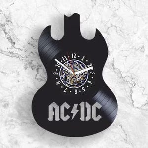May include: A black wall clock shaped like an electric guitar, crafted from a vinyl record. The clock face features white numbers from 1 to 12, white hands, and a visible gear mechanism in the center. The AC/DC logo is printed in white at the base.