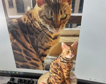 May include: A detailed image of a Bengal cat figurine with a matching image in the background. The figurine is brown and black with a sitting pose. The cat's eyes are green. The figurine is on a round, light-colored base.