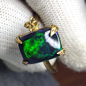 Könnte beinhalten: Ein goldener Ring mit einem großen, quadratisch geschliffenen Opal mit lebendigen grünen und blauen Farben. Der Opal ist in einem strukturierten Goldband mit floralen und krallenartigen Akzenten gefasst, was ein einzigartiges Design ergibt. Ein auffälliges Schmuckstück.