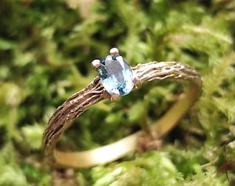 May include: A gold ring with a light blue oval gemstone. The band has a textured, organic design, resembling tree bark. The ring is set against a blurred green background, highlighting the jewelry's details.