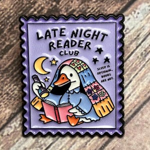 Peut inclure: Une broche en émail rectangulaire en forme de timbre-poste sur fond violet clair. Elle représente un canard blanc sous une courtepointe colorée, tenant une lampe de poche et un livre rouge. Texte : LATE NIGHT READER CLUB et SLEEP IS OPTIONAL, BOOKS ARE NOT.