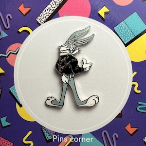 May include: A Bugs Bunny enamel pin in a walking pose, giving a thumbs up. The pin features Bugs Bunny in a black jacket with the words "Bugs Bunny" on the back. The pin is on a white circle, with a purple background.