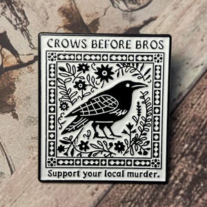 Op de afbeelding: Zwart-witte emaille pin met de tekst "CROWS BEFORE BROS" en "Support your local murder." De pin toont een zwarte kraaienillustratie omringd door bloemen en geometrische ontwerpen. Een decoratief accessoire.