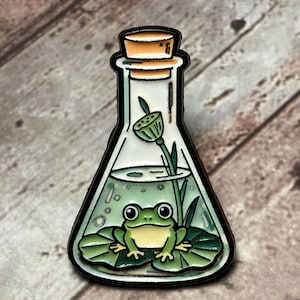 Peut inclure: Une broche en émail triangulaire en forme de fiole de laboratoire contenant une grenouille verte assise sur un nénuphar dans l'eau. Une gousse de lotus sur une tige émerge de l'eau. La fiole est scellée par un bouchon en liège et un contour noir.