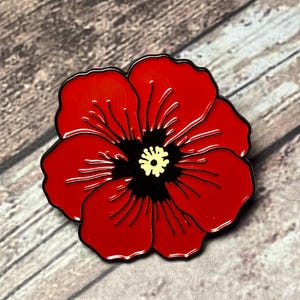Puede incluir: Un broche de flor de amapola roja con contorno y centro negros. El centro de la flor es negro con un acento amarillo. La amapola es un símbolo de recuerdo y se usa para conmemorar a los veteranos.