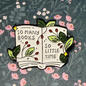Peut inclure: Épinglette en émail blanc en forme de livre ouvert avec le texte "SO MANY BOOKS SO LITTLE TIME". La broche présente des feuilles vertes, des baies rouges et des étoiles jaunes. Le fond est un motif floral.