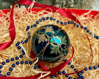 May include: A teal Christmas ornament with gold floral detailing rests in a decorative box filled with straw. A red ribbon and blue beaded garland surround the ornament. The box has a red and white striped pattern.