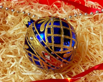 May include: A blue and gold Christmas ornament with a grid pattern and jewel accents rests in a box filled with straw. Red ribbon and silver beads surround the ornament, creating a festive holiday scene.