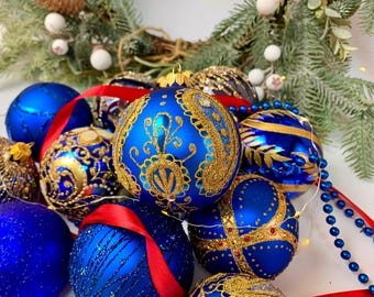 May include: A collection of blue Christmas ornaments with gold and glitter accents. Some ornaments have intricate gold patterns, while others feature stripes or cross designs. Red ribbon and blue beaded garland add festive touches.
