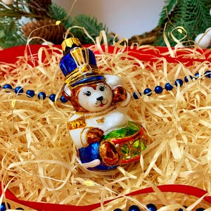 Puede incluir: Adorno navideño festivo con forma de mono baterista, vestido con uniforme azul y dorado. El adorno es blanco, marrón y verde, y está colocado en un lecho de paja con una cinta roja y cuentas azules. Una decoración navideña.