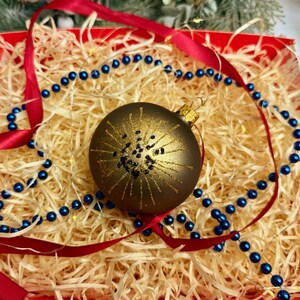 Puede incluir: Una bola decorativa de Navidad con un diseño dorado y negro, colocada en una caja roja llena de paja. Una cinta roja y una guirnalda de cuentas azules completan la exhibición festiva.