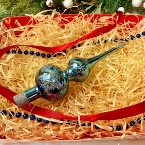 May include: A blue and silver Christmas tree topper with a snowflake design rests in a red box filled with straw. A red ribbon and blue beaded garland decorate the box, suggesting a festive holiday theme.